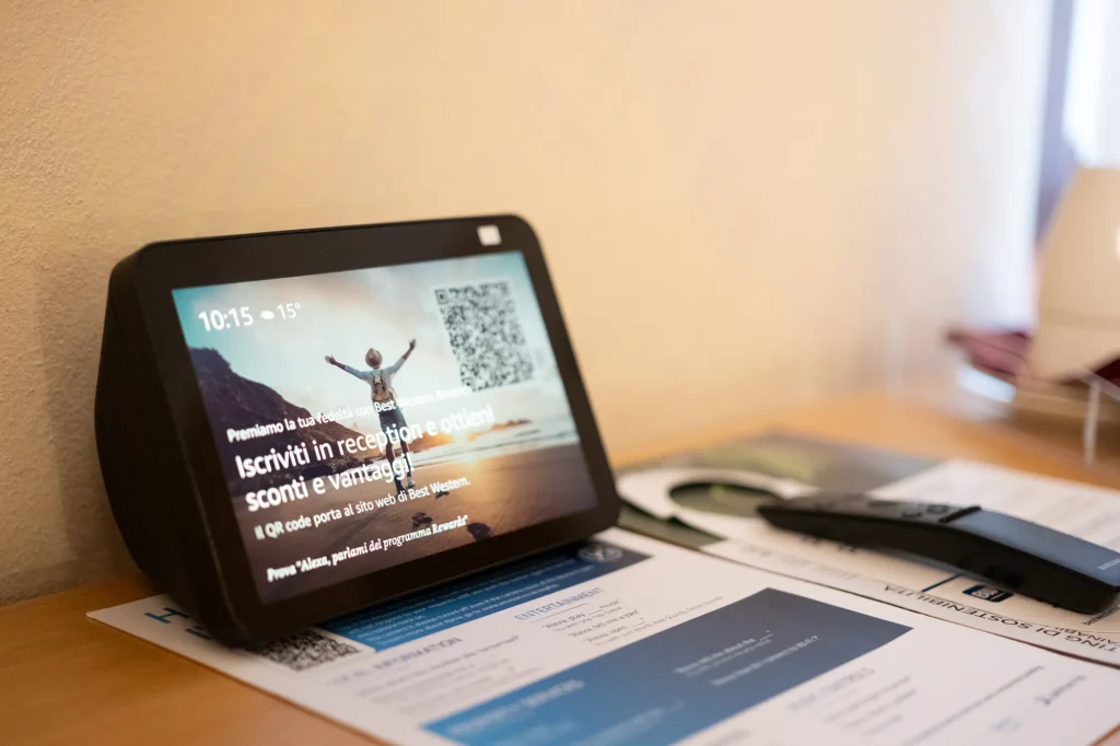 Schermo digitale appoggiato su un tavolo che mostra un messaggio informativo con testo promozionale e codici QR, affiancato da telecomandi e fogli stampati con informazioni