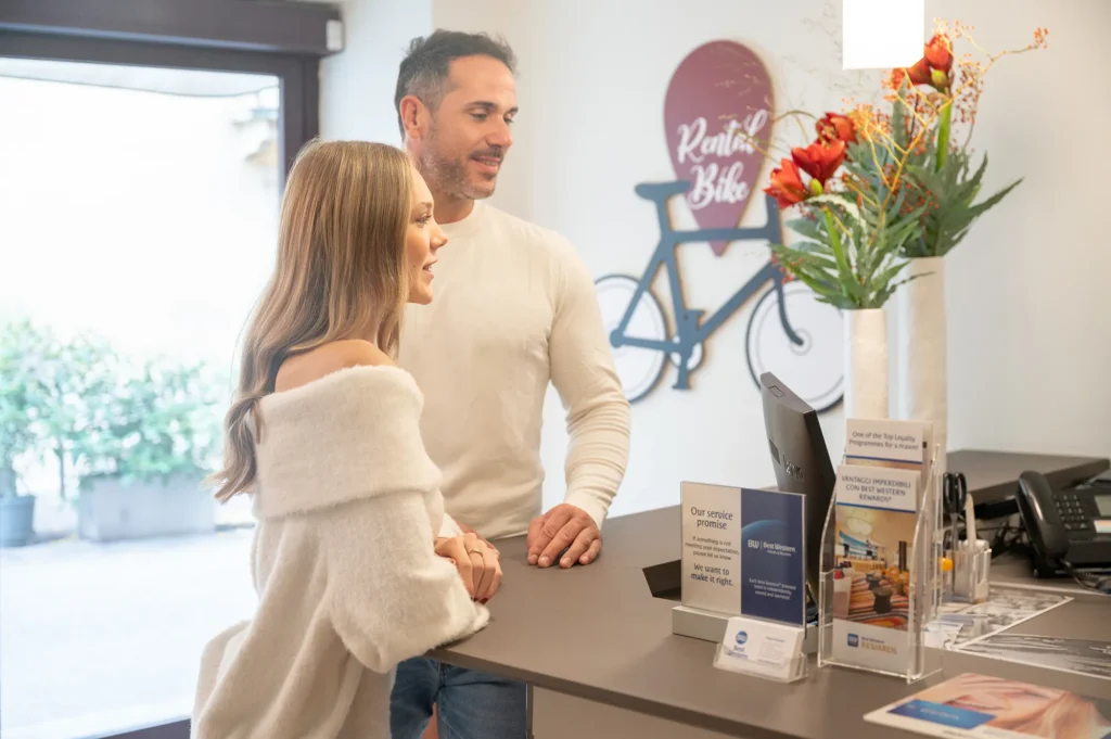 Due persone alla reception dell’hotel parlano con un addetto, davanti a un banco con brochure informative e un’insegna dedicata al servizio di noleggio biciclette