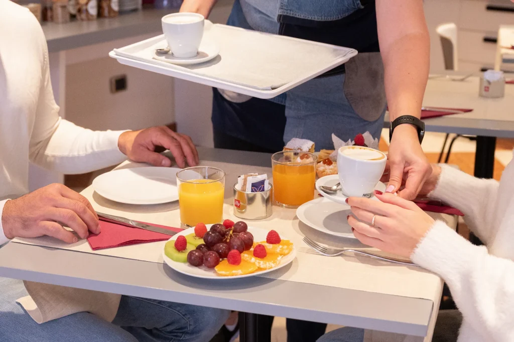Two people seated at a breakfast table with fruit plates and glasses of orange juice, while a server places a cup of coffee on the table
