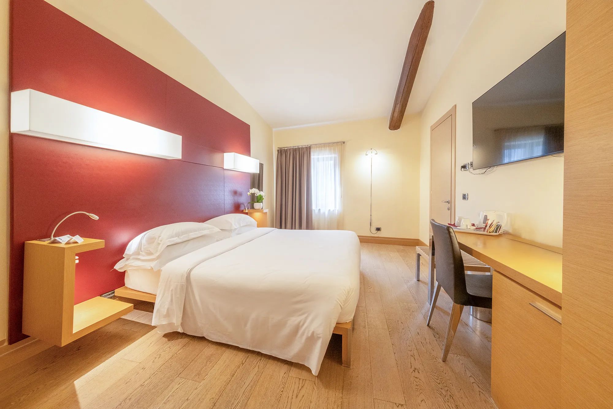 Camera d’albergo con letto matrimoniale, testata rossa illuminata, finestra con tende, scrivania con sedia e lampada, grande televisore a parete e trave in legno sul soffitto