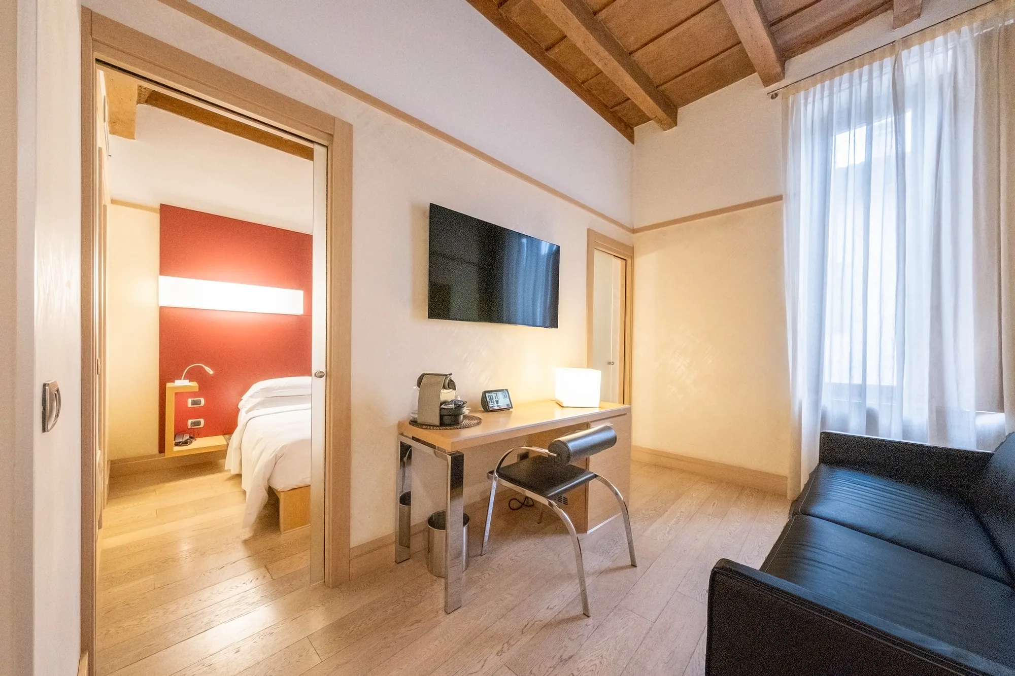 Area soggiorno di una camera d’albergo con divano scuro, scrivania in legno con macchina da caffè e lampada, sedia nera, televisore a parete e porta scorrevole aperta che mostra la camera da letto con testata rossa illuminata