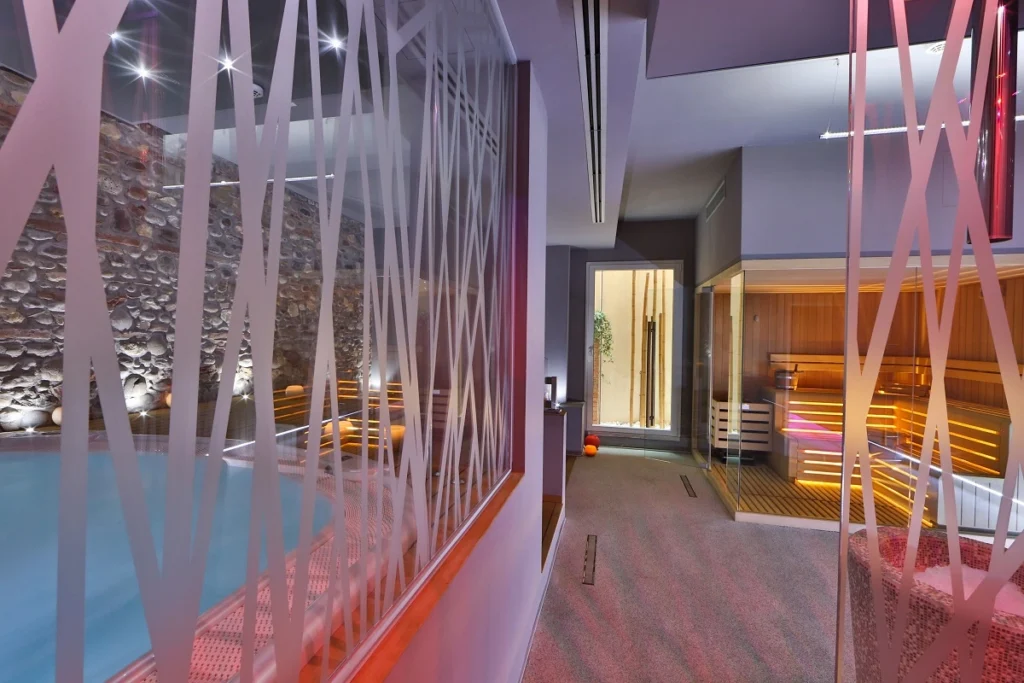 Wellness area with indoor pool and sauna illuminated by colored lights, in a modern setting with glass and stone walls