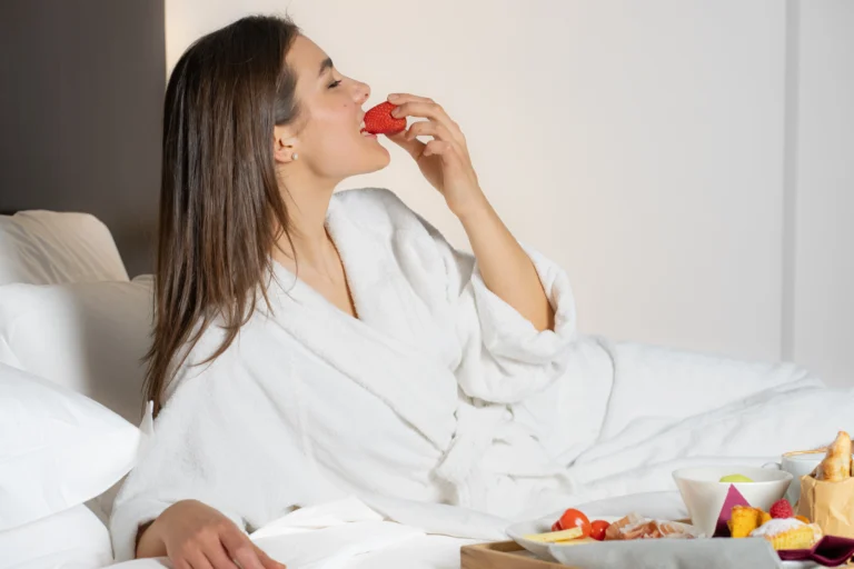 Donna in accappatoio bianco seduta sul letto mentre mangia una fragola da un vassoio con colazione.
